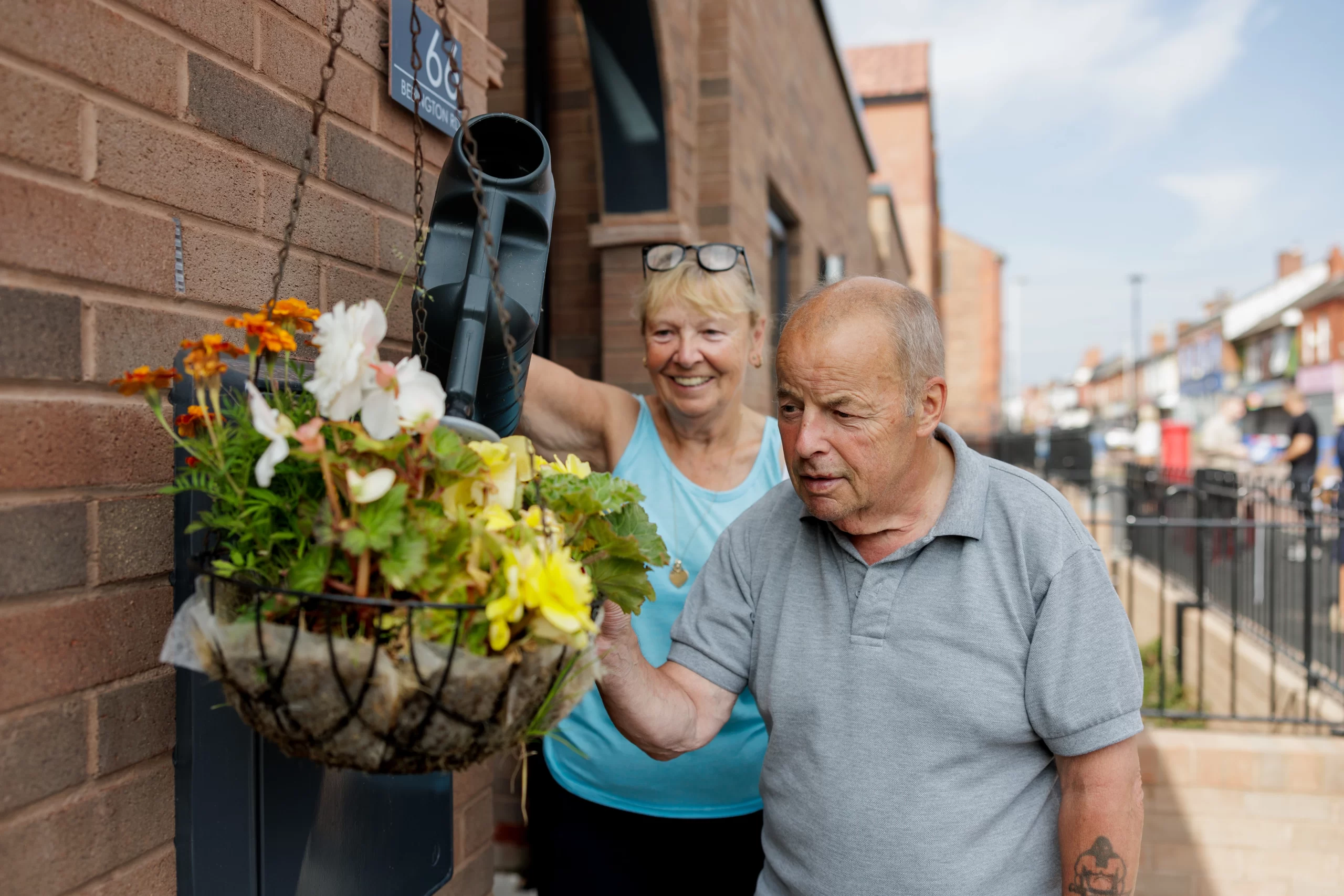 lady in a blue vest watering a hanging basket stood next to gentleman in grey polo t shirt